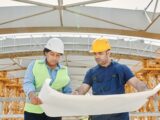 Dois engenheiros com capacete e colete analisam a planta de um projeto em um canteiro de obras