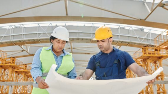 Dois engenheiros com capacete e colete analisam a planta de um projeto em um canteiro de obras