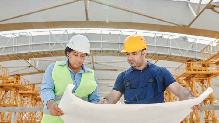 Dois engenheiros com capacete e colete analisam a planta de um projeto em um canteiro de obras